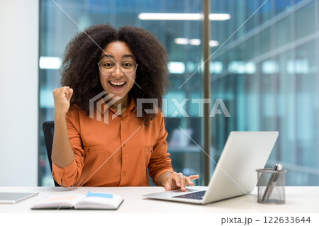 Enthusiastic African American businesswoman celebrating achievement with laptop in modern office. Expresses success, motivation, accomplishment, and confidence in workspace environment. 122633644