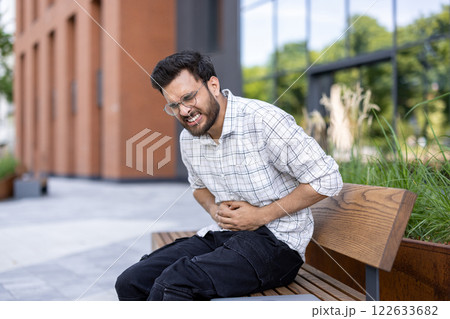 Contorted from severe pain, a young Indian man sits on a bench in the street and holds his stomach with his hands. 122633682