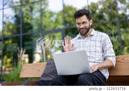 Smiling man using laptop outdoors holding video call with headphones wearing casual attire. Background of modern building , emphasizing connection, technology, and remote communication Smiling man using laptop outdoors holding video call with headphones wearing casual attire. Background of modern building , emphasizing connection, technology, and remote communication 122633716