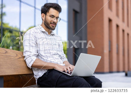 Young man sitting on bench using laptop. Outdoor setting near modern building, focusing on work or study. Warm and productive atmosphere. 122633735