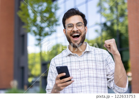 Close-up portrait of a young smiling Indian man standing outside on the street, holding a mobile phone in his hand and rejoicing in success celebrating a victory for the camera. 122633753