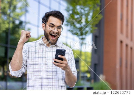 Photo of a happy young Indian man standing outside on the street, looking at the phone screen and rejoicing in success, raising his hand up. 122633773