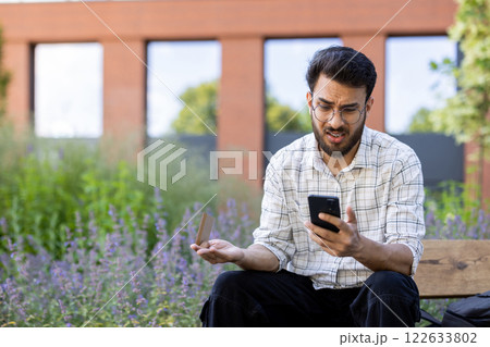 Worried and frustrated young indian man wearing glasses sitting outside on a bench and looking upset at the mobile phone ,screen while waving his hand incoherently. Worried and frustrated young indian man wearing glasses sitting outside on a bench and looking upset at the mobile phone ,screen while waving his hand incoherently. 122633802