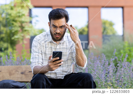 Man sitting on bench in park checking smartphone with worried expression. Urban area in background with greenery. Concept of stress, technology, communication. 122633803