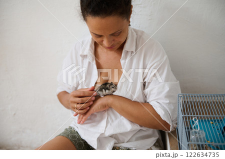 Woman holding a hamster inside her shirt while sitting by a pet's cage Woman holding a hamster inside her shirt while sitting by a pet's cage 122634735