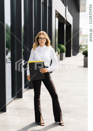 Elegant confident young woman, formally dressed, stands outdoors against the background of the business center, holding a folder with documents, looks at camera Elegant confident young woman, formally dressed, stands outdoors against the background of the business center, holding a folder with documents, looks at camera 122635450