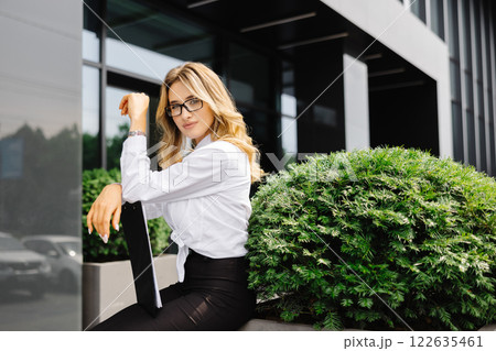 Elegant confident young woman, formally dressed, stands outdoors against the background of the business center, holding a folder with documents, looks at camera 122635461