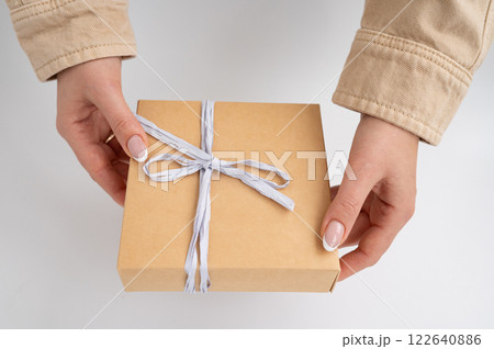 Caucasian female hands holding brown gift box with white ribbon. National Cartonnage Day Caucasian female hands holding brown gift box with white ribbon. National Cartonnage Day 122640886