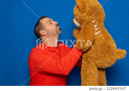 Bearded hispanic man in his 40s wearing a red sweater holding up a teddy bear while blowing kisses and cuddling at it, isolated on blue studio background. Bearded hispanic man in his 40s wearing a red sweater holding up a teddy bear while blowing kisses and cuddling at it, isolated on blue studio background. 122643547