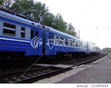 A blue passenger train rests at a quiet rural station surrounded by trees in the early morning light. The platform is empty, showcasing the serene atmosphere of the moment. 122643954
