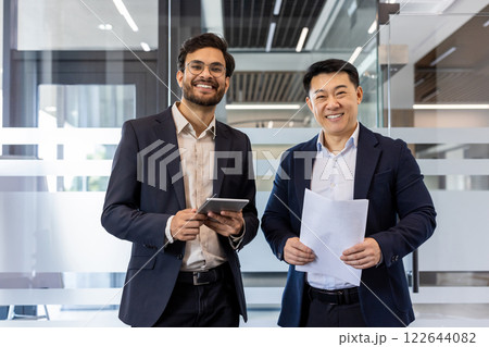 Smiling businessmen standing in modern office showcasing teamwork and professionalism. One holding tablet, other documents, reflecting success and collaboration in business environment. Smiling businessmen standing in modern office showcasing teamwork and professionalism. One holding tablet, other documents, reflecting success and collaboration in business environment. 122644082