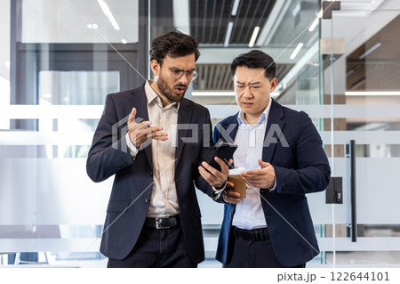 Two businessmen in modern office setting intensely discuss news displayed on smartphone. Colleagues in professional attire express concern, highlighting teamwork and urgency in business communication Two businessmen in modern office setting intensely discuss news displayed on smartphone. Colleagues in professional attire express concern, highlighting teamwork and urgency in business communication 122644101