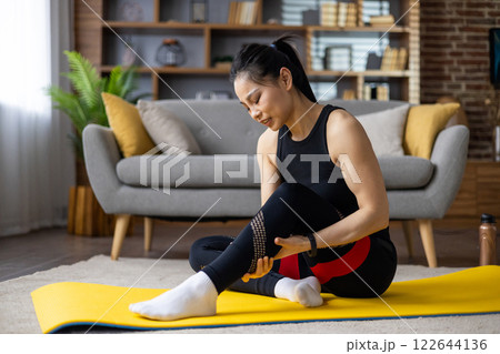 Asian woman injured leg during workout at home, seated on yoga mat, holding leg in pain. Dressed in athletic wear, captures moment of discomfort, emphasizing fitness, health, and injury concept. 122644136