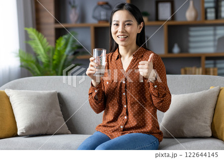 Asian woman smiling on sofa holding glass of water while giving thumbs up. Wearing casual brown polka dot shirt, jeans, radiating positivity, relaxation, well-being, hydration, healthy lifestyle. Asian woman smiling on sofa holding glass of water while giving thumbs up. Wearing casual brown polka dot shirt, jeans, radiating positivity, relaxation, well-being, hydration, healthy lifestyle. 122644145