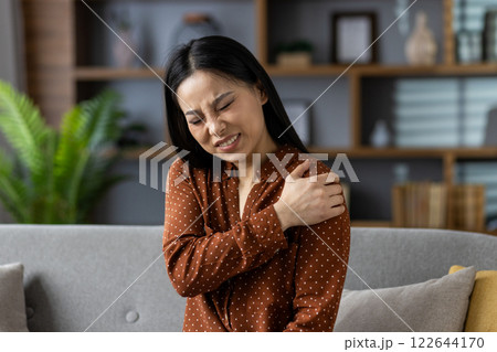 Asian woman sitting on sofa with shoulder pain, seeking relief. Expression shows discomfort, wearing polka dot blouse in modern living room setting. Concept of physical discomfort. Asian woman sitting on sofa with shoulder pain, seeking relief. Expression shows discomfort, wearing polka dot blouse in modern living room setting. Concept of physical discomfort. 122644170