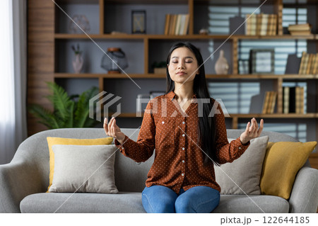 Asian woman practicing meditation on sofa, eyes closed, embracing calm and mindfulness. Dressed casually, sitting in relaxed pose. Living room setting enhances peaceful and comfortable atmosphere. Asian woman practicing meditation on sofa, eyes closed, embracing calm and mindfulness. Dressed casually, sitting in relaxed pose. Living room setting enhances peaceful and comfortable atmosphere. 122644185