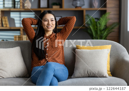 Smiling woman sitting on sofa, feeling relaxed and happy. Hands behind head, exuding peace and contentment. Dressed casually in polka dot shirt and jeans. Modern home atmosphere. Smiling woman sitting on sofa, feeling relaxed and happy. Hands behind head, exuding peace and contentment. Dressed casually in polka dot shirt and jeans. Modern home atmosphere. 122644187