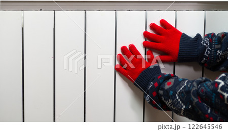 Hands in gloves on a radiator. Central heating radiator at home. A child in woolen gloves warms his hands near the heater Hands in gloves on a radiator. Central heating radiator at home. A child in woolen gloves warms his hands near the heater 122645546