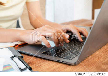 Hands typing on a laptop in a comfortable workspace, featuring a notepad and pen on a wooden desk. Hands typing on a laptop in a comfortable workspace, featuring a notepad and pen on a wooden desk. 122649513