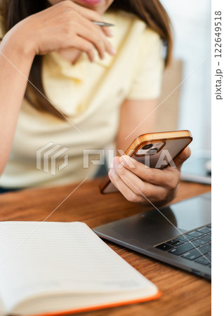 Woman holding smartphone at desk with laptop and notebook, representing modern technology and communication in a casual work setting. 122649518
