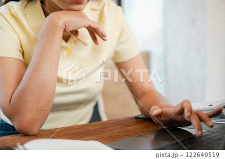 A woman in a yellow shirt multitasking with a laptop and smartphone at a wooden table, showcasing modern technology use in a casual home environment. A woman in a yellow shirt multitasking with a laptop and smartphone at a wooden table, showcasing modern technology use in a casual home environment. 122649519