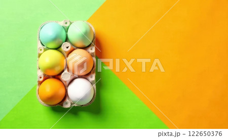 A carton filled with six beautifully colored eggs is placed on a bright green and orange background. The eggs feature pastel shades and a glossy finish, showcasing creativity and festivity. 122650376
