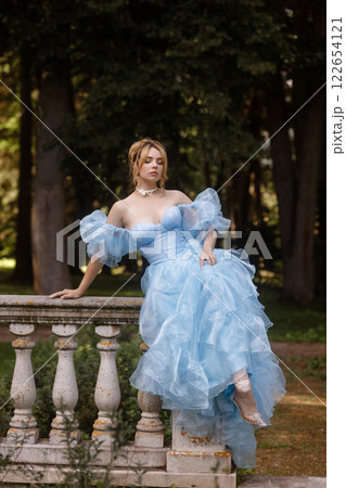 Romantic Woman in a Light Blue Gown Posing on a stone balustrade in a picturesque garden. 122654121