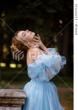 Romantic Woman in a Light Blue Gown Posing on a stone balustrade in a picturesque garden. 122654122
