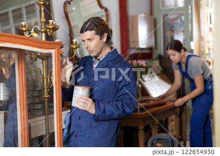 Skilled middle-aged male furniture repair specialist in uniform restoring and painting cupboard furniture during workday in workshop Skilled middle-aged male furniture repair specialist in uniform restoring and painting cupboard furniture during workday in workshop 122654930