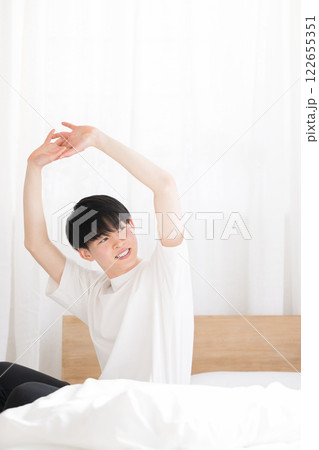 朝日を浴びるベッドで寝起きの若い男性 腕を伸ばすストレッチをする 右にコピースペースあり 朝日を浴びるベッドで寝起きの若い男性 腕を伸ばすストレッチをする 右にコピースペースあり 122655351