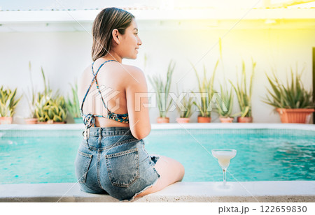 Girl sitting on the edge of the pool with tropical drink. Rear view of girl sitting on the edge of the pool with a drink. Woman on vacation sitting on the edge of swimming pool with a drink Girl sitting on the edge of the pool with tropical drink. Rear view of girl sitting on the edge of the pool with a drink. Woman on vacation sitting on the edge of swimming pool with a drink 122659830
