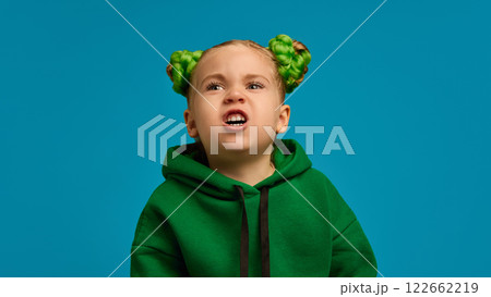 Portrait of emotive 5 years old girl in comfortable green hoodie making faces, grimacing against blue studio background 122662219