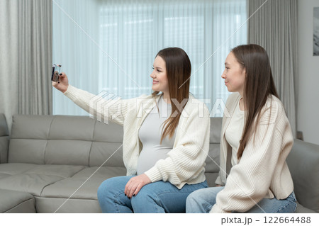 Young pregnant woman taking a selfie with a friend at home on a sofa, smiling, with copy space. Concept of friendship, maternity, capturing special memories during pregnancy. 122664488