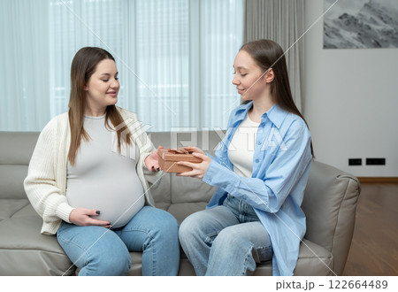 Young woman giving a gift for baby to her pregnant friend while sitting on a couch and smiling in a cozy home. Concept of maternity, celebration, friendship, anticipation, and emotional connection. 122664489