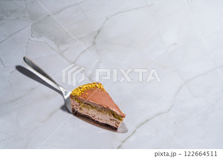 Slice of Dubai chocolate cheesecake with pistachio paste and kataifi covered in chocolate ganache on a cake spatula. Marble background with copy space. Indulgence, gourmet desserts, culinary 122664511