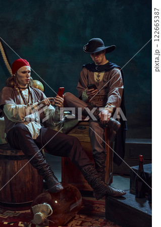 Young men, medieval pirates in vintage costumes sitting with smartphones, scrolling social media feed, discussing news against dark background in dimly-lit room Young men, medieval pirates in vintage costumes sitting with smartphones, scrolling social media feed, discussing news against dark background in dimly-lit room 122665387