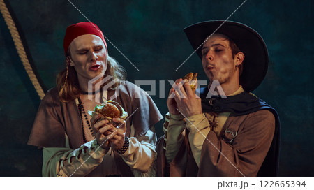 Close-up portrait of medieval pirates in hat and bandana, passionately eating burgers against dark vintage background. Close-up portrait of medieval pirates in hat and bandana, passionately eating burgers against dark vintage background. 122665394