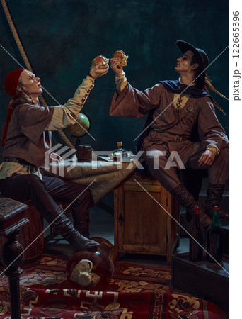 Two pirates, one in red bandana and other in feathered hat, raising burgers, enjoy fast food in dimly lit room with vintage maritime props Two pirates, one in red bandana and other in feathered hat, raising burgers, enjoy fast food in dimly lit room with vintage maritime props 122665396