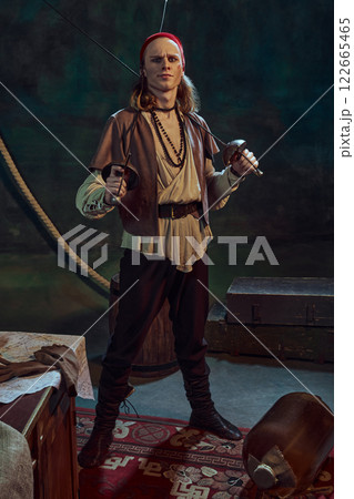 Young man wish wounded face, pirate standing with crossed swords, with playful smirk on his face against dark vintage background 122665465