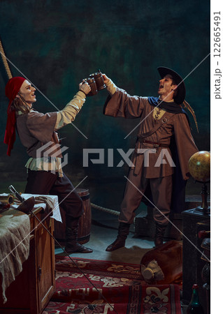 Two pirates, in red bandana and in feathered hat, clinging wooden mugs filled with beer, enjoy drink in dimly lit room with vintage maritime props 122665491