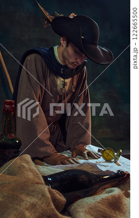 Focused man pirate captain in feathered hat and gloves, looking at vintage map and compass, against dark background with maritime props and bottles. 122665500