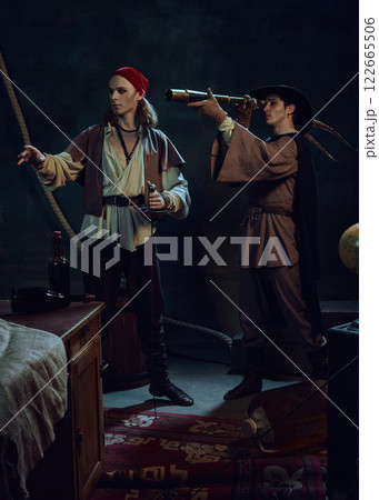 Two pirates standing in dim candlelight, one peering through spyglass, other gripping sword. Two pirates standing in dim candlelight, one peering through spyglass, other gripping sword. 122665506