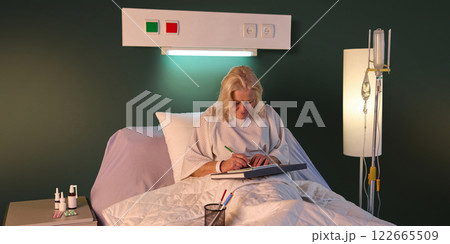 Elderly woman in hospital gown sitting in bed in her ward in the evening and drawing against green wall and drip equipment. 122665509