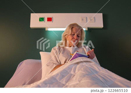 Elderly woman in hospital gown sitting in bed in her ward in the evening and making notes in her journal. Planning, writing down thoughts Elderly woman in hospital gown sitting in bed in her ward in the evening and making notes in her journal. Planning, writing down thoughts 122665510