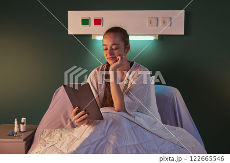 Young woman sitting in bed in gown in the evening and using tablet for communication, education, books reading. Modern clinic ward Young woman sitting in bed in gown in the evening and using tablet for communication, education, books reading. Modern clinic ward 122665546