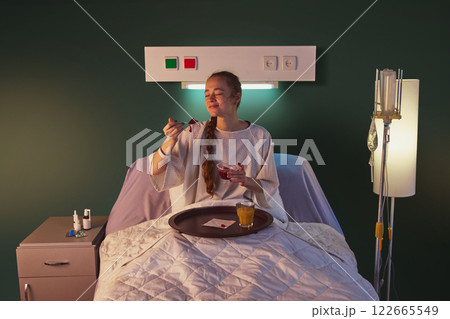 Young red-haired woman in her 20s, patient in white hospital gown, sitting in bed, and eating jelly for dinner with orange juice Young red-haired woman in her 20s, patient in white hospital gown, sitting in bed, and eating jelly for dinner with orange juice 122665549