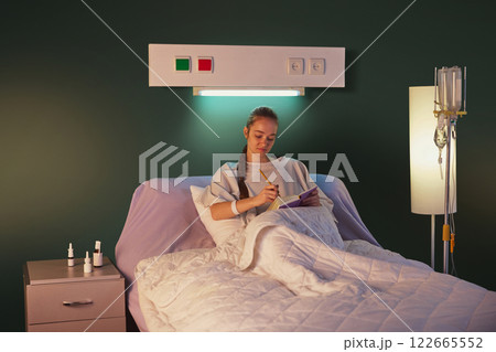Young woman with braid, in hospital gown, lying in bed with drip equipment standing near and making notes on her journal against green wall 122665552