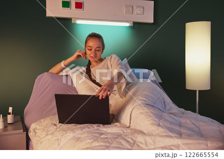 Young woman in her 20s lying on bed in hospital looking at laptop against green wall with lightning on and meds on bed table 122665554