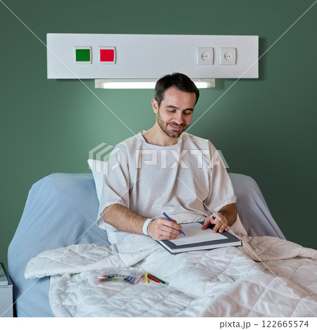 Man in hospital gown sitting in bed and drawing with colorful pencils, leisure time during recovery process in modern clinic 122665574