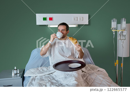Man in his 40s sitting in hospital bed in gown and having breakfast, drinking tea with croissant against green wall, drip equipment, and meds on table 122665577